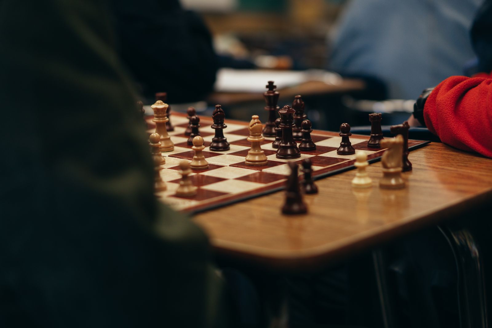 A wooden chess set mid-game on a table, with players' arms resting beside it at one of many chess tournaments