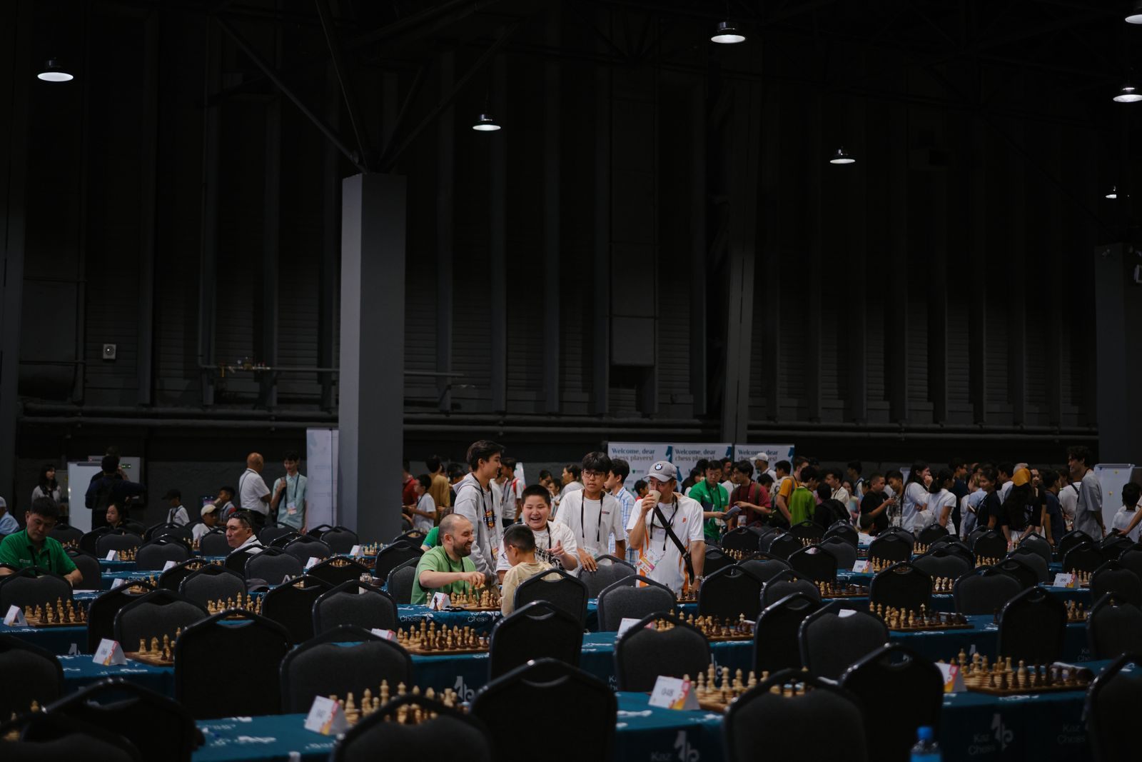 Rows of players at chess boards in a large, darkened hall during a mind sports tournament