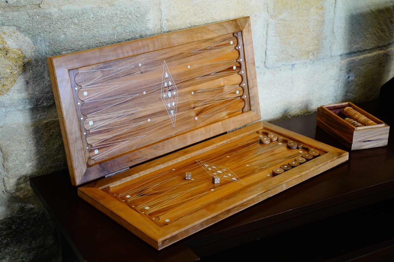 An open wooden backgammon board with checkers and dice on a table against a stone wall